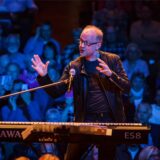 Der Komponist und Produzent Dieter Falk sitzt auf einer Bühne an einem Keyboard, hinter ihm ist das Publikum zu sehen.