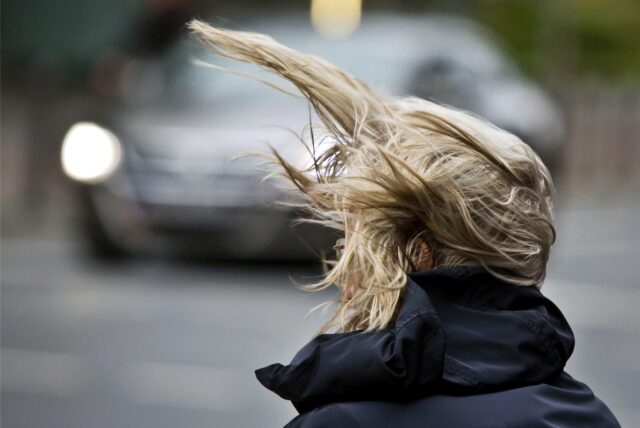 Eine Frau steht an einer Kreuzung, während starker Wind ihre Frisur durcheinander bringt.