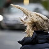 Eine Frau steht an einer Kreuzung, während starker Wind ihre Frisur durcheinander bringt.
