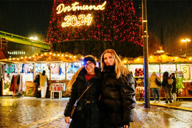 Zwei Personen posieren auf dem Dortmunder Weihnachtsmarkt. Hinter ihnen steht der 45 Meter hohe Weihnachtsbaum mit der Aufschrift Dortmund 2024.