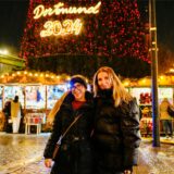 Zwei Personen posieren auf dem Dortmunder Weihnachtsmarkt. Hinter ihnen steht der 45 Meter hohe Weihnachtsbaum mit der Aufschrift Dortmund 2024.