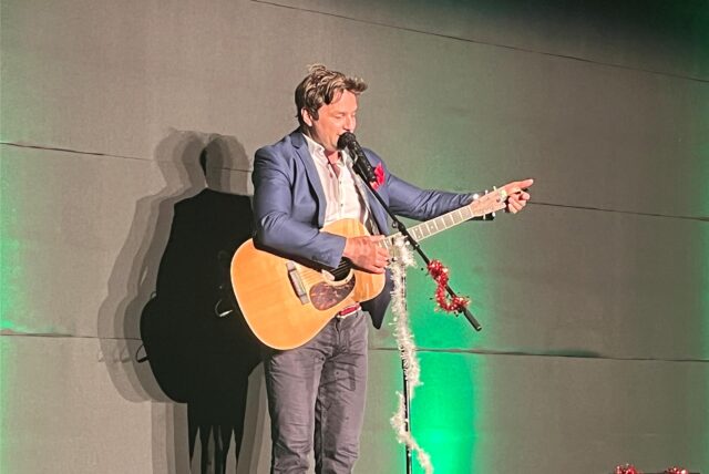 Ein Mann mit Gitarre vor einem heruntergelassenen Vorhang.