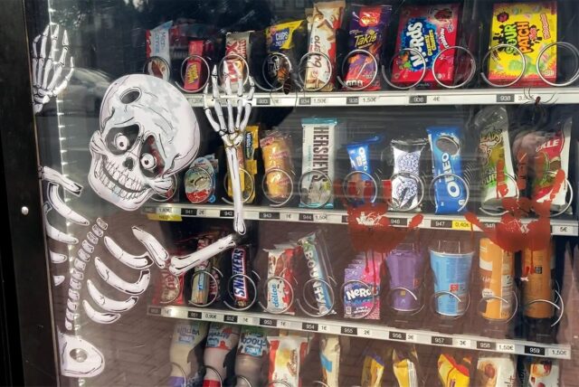 Verschiedene Süßigkeiten und Getränke liegen in der Auslage eines Süßwarenautomaten. An der Scheibe klebt ein Aufkleber in Form eines Skeletts.