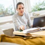 Eine Frau sitzt an einem Tisch, auf dem ein Laptop, ein Smartphone und ein Aktenordner liegen.