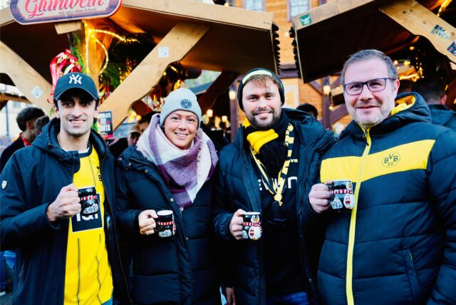 Vier BVB-Fans stehen vor einer Glühweinhütte auf dem Dortmunder Weihnachtsmarkt und trinken aus einer Pinguin-Tasse.