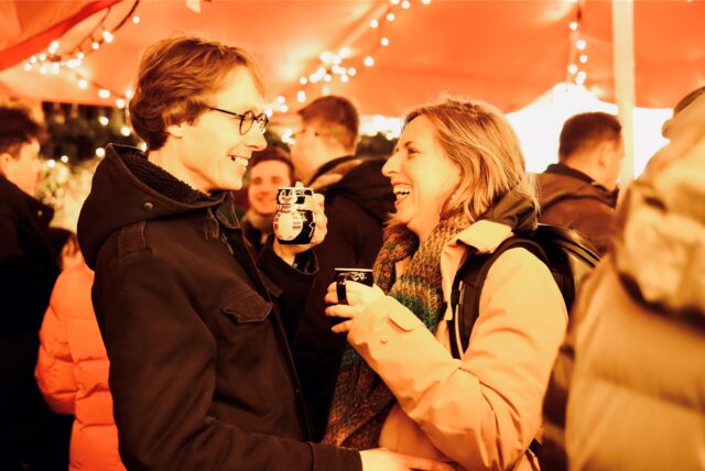 Ein Mann und eine Frau, die unter einem leuchtenden Zelt auf dem Dortmunder Weihnachtsmarkt stehen, lächeln sich an. Sie haben die Pinguin-Weihnachtsmarkttasse in der Hand.
