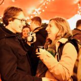 Ein Mann und eine Frau, die unter einem leuchtenden Zelt auf dem Dortmunder Weihnachtsmarkt stehen, lächeln sich an. Sie haben die Pinguin-Weihnachtsmarkttasse in der Hand.