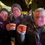 Zwei Frauen und ein Mann halten auf dem 36. Holzwickeder Weihnachtsmarkt ihre Tassen hoch und lächeln in die Kamera.