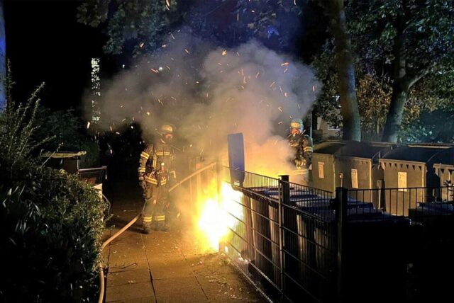 Ein Feuerwehrmann steht vor brennenden Mülltonnen.