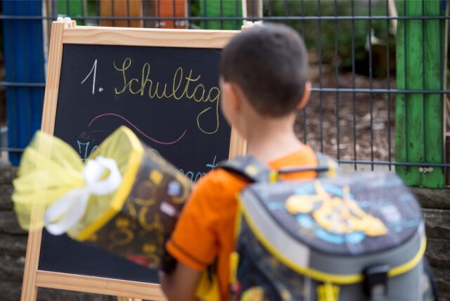 „1. Schultag“ steht auf einer Tafel, vor der ein Erstklässler steht.