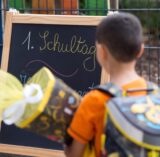 „1. Schultag“ steht auf einer Tafel, vor der ein Erstklässler steht.