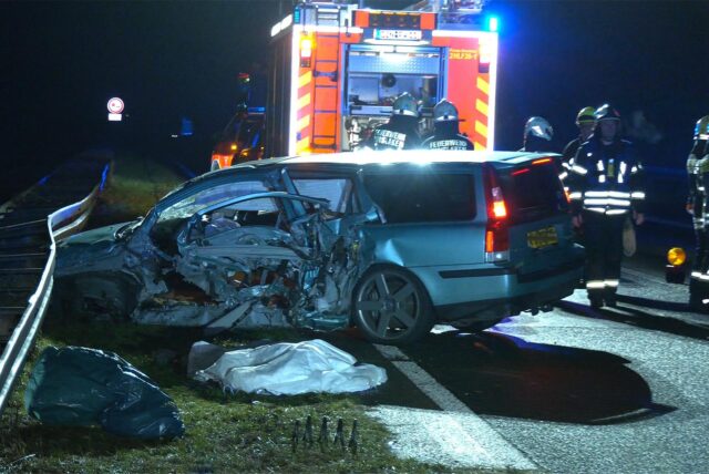 Das zerstörte Auto steht frontal an einer Leitplanke. Dahinter sind Rettungswagen und Rettungskräfte zu sehen.