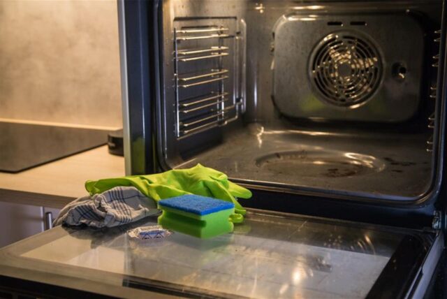 Vor einem geöffneten Backofen liegen ein Schwamm, Handschuhe und ein Spülmaschinen-Tab.