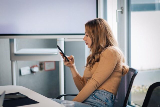 Eine Frau sitzt im Büro und hat ein Handy in der Hand.
