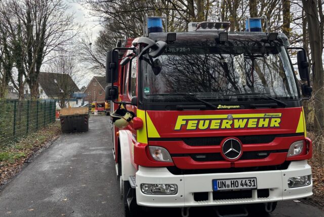 Ein Einsatzwagen der Feuerwehr steht auf einer Straße vor dem Wertstoffhof in Holzwickede. Im Hintergund ist ein Container zu sehen.