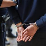Zu sehen sind zwei Personen von hinten – einer von ihnen hat die Hände auf dem Rücken in Handschellen und wird vom anderen geführt