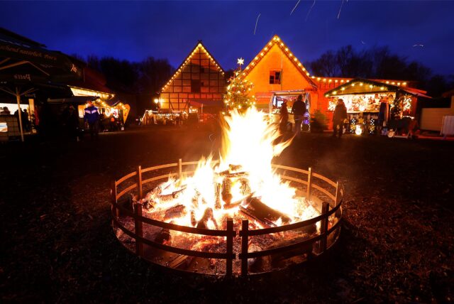 Ein Lagerfeuer sorgt im Dunkeln für ein besonderes Flair auf dem Weihnachtsmarkt auf Gut Keinemann in Rünthe. Im Hintergrund sind beleuchtete Häuser und Buden zu sehen.