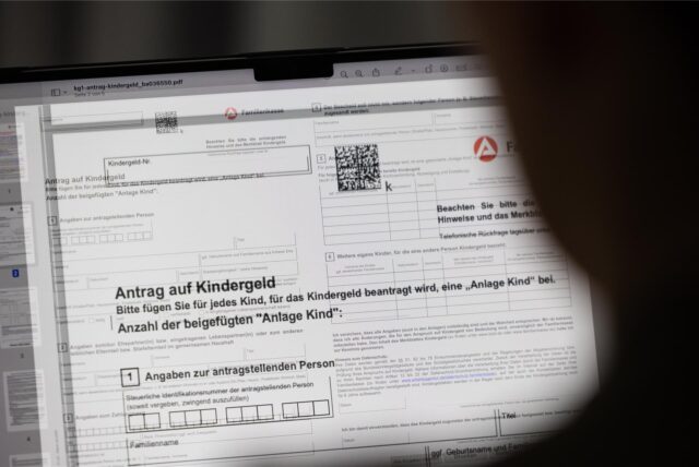 Auf einem Computerbildschirm ist der Ausschnitt eines Antrags auf Kindergeld zu sehen. Im Vordergrund erscheint am rechten Bildrand die unscharfe, dunkle Form einer Person, die auf den Bildschirm schaut. Von der Person sind der halbe Kopf und der Hals zu sehen.