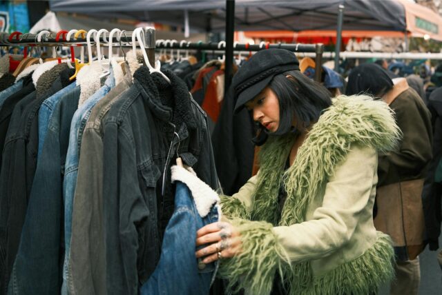 Auf einem Trödelmarkt durchstöbert eine Frau in einer grünen Jacke und einer schwarzen Kappen einen Stand mit verschiedenen Jacken – sie hält eine gefütterte Jeansjacke in der Hand.