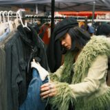 Auf einem Trödelmarkt durchstöbert eine Frau in einer grünen Jacke und einer schwarzen Kappen einen Stand mit verschiedenen Jacken – sie hält eine gefütterte Jeansjacke in der Hand.