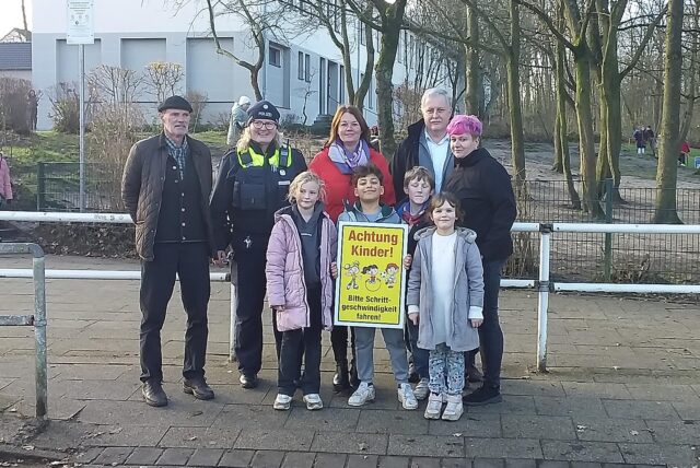 Eine Gruppe Menschen steht vor einem weißen Zaun, alle lächeln in die Kamera. Ein Junge hält das Schild „Achtung Kinder“ hoch.