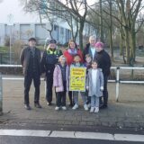 Eine Gruppe Menschen steht vor einem weißen Zaun, alle lächeln in die Kamera. Ein Junge hält das Schild „Achtung Kinder“ hoch.