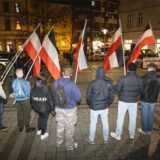 Eine Gruppe junger Erwachsener in Winterkleidung, teils mit Kapuzen, steht auf einem gepflasterten Platz in einem Halbkreis. Sie stehen mit dem Rücken zur Kamera, sechs von ihnen