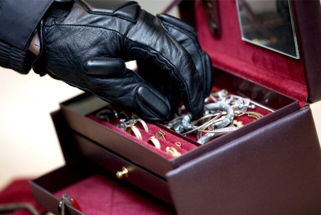 Eine Hand mit schwarzem Handschuh greift in eine dunkelrote Schmuckschatulle und greift sich Schmuck.