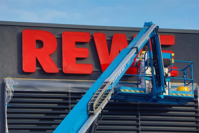 Ein Bild von einem Hubkran. Darauf sind Mitarbeiter zu sehen die an dem großen Logo des Supermarkts arbeiten.