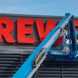 Ein Bild von einem Hubkran. Darauf sind Mitarbeiter zu sehen die an dem großen Logo des Supermarkts arbeiten.