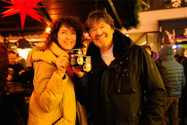 Eine lockige Frau und ein Mann mit Brille stoßen auf dem Dortmunder Weihnachtsmarkt mit der Pinguin-Tasse an.