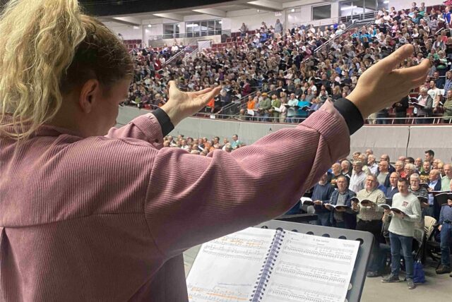 Eine Frau dirigiert eine Halle voller Menschen beim Singen.