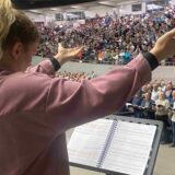 Eine Frau dirigiert eine Halle voller Menschen beim Singen.