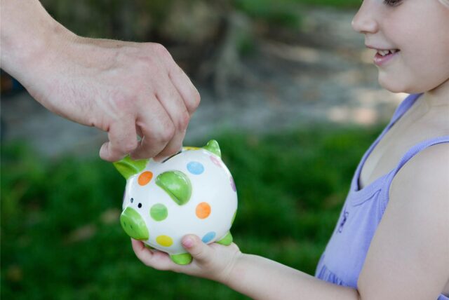 Ein Kind hält ein Sparschwein in der Hand. Eine erwachsene Hand wirft eine Münze hinein.