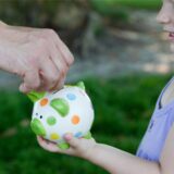 Ein Kind hält ein Sparschwein in der Hand. Eine erwachsene Hand wirft eine Münze hinein.