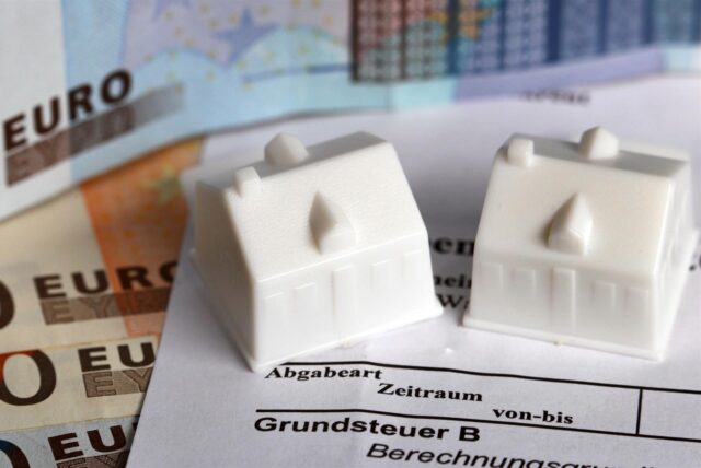 Zwei Spielhäuschen stehen auf einem Grundsteuerbescheid. Darunter liegen mehrere Geldscheine.