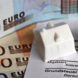 Zwei Spielhäuschen stehen auf einem Grundsteuerbescheid. Darunter liegen mehrere Geldscheine.