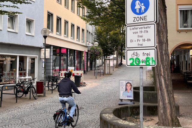 Ein Radfahrer fährt in Unnas Fußgängerzone hinein. In der Bildmontage sind Uhrzeiten auf einem Verkehrsschild grün übermalt.