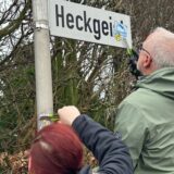 Eine Frau und ein Mann kratzen mit Messern Aufkleber von einem Straßenschild.