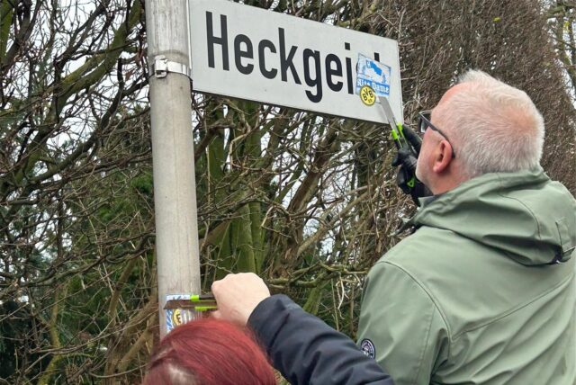 Eine Frau und ein Mann kratzen mit Messern Aufkleber von einem Straßenschild.