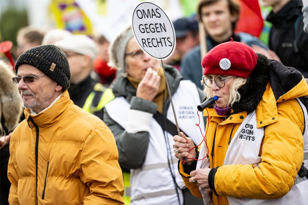 In einer Demonstrationsmenge stehen im Vordergrund zwei ältere Personen mit gelben Jacken. Auf einem Schild steht Omas gegen Rechts. In einer Demonstrationsmenge stehen im Vordergrund zwei ältere Personen mit gelben Jacken. Auf einem Schild steht Omas gegen Rechts.