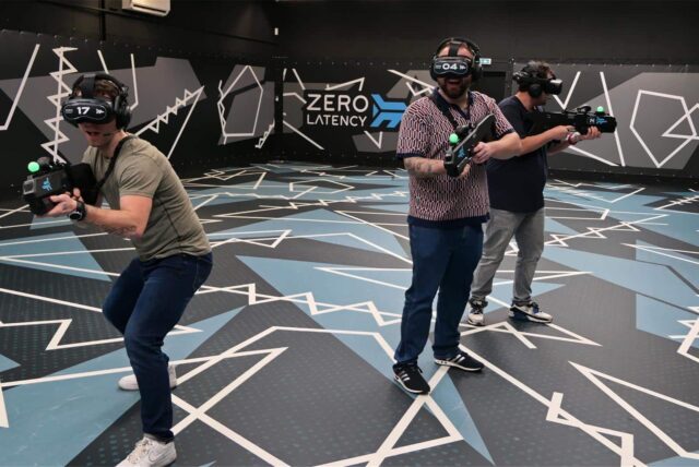 Drei Spieler mit VR-Brillen und Spielzeuggewehren stehen auf einer leeren Spielfläche von Zero Latency.