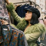Eine Frau mit grüner Jacke und schwarzer Mütze durchstöbert einen Stand mit Hemden und Jacken auf einem Trödelmarkt.