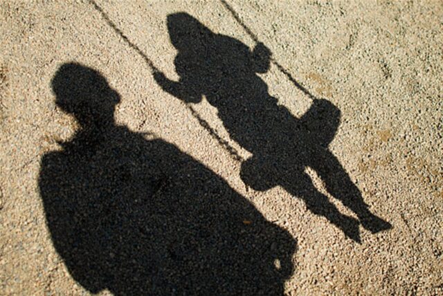 Der Schatten von einem Mann und einem schaukelnden Kind fallen auf Sand auf einem Spielplatz in Braunschweig. (Archivbild)