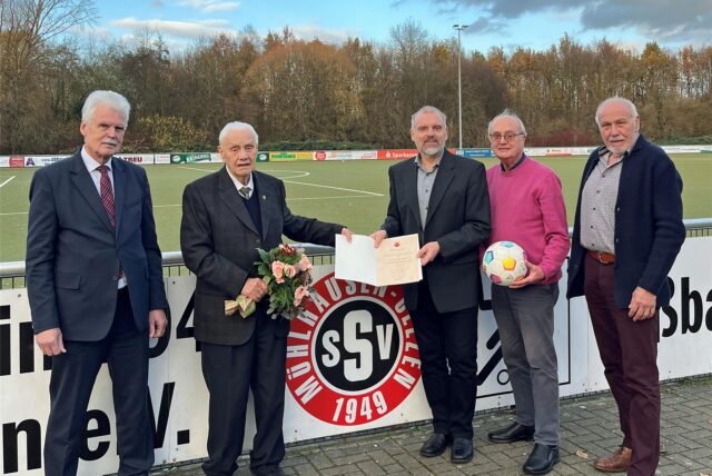 Fünf Männer stehen an der Bande des Sportplatzes des SSV Mühlhausen. Einer hält einen Blumenstrauß, einer eine Urkunde, einer einen Ball.