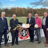 Fünf Männer stehen an der Bande des Sportplatzes des SSV Mühlhausen. Einer hält einen Blumenstrauß, einer eine Urkunde, einer einen Ball.