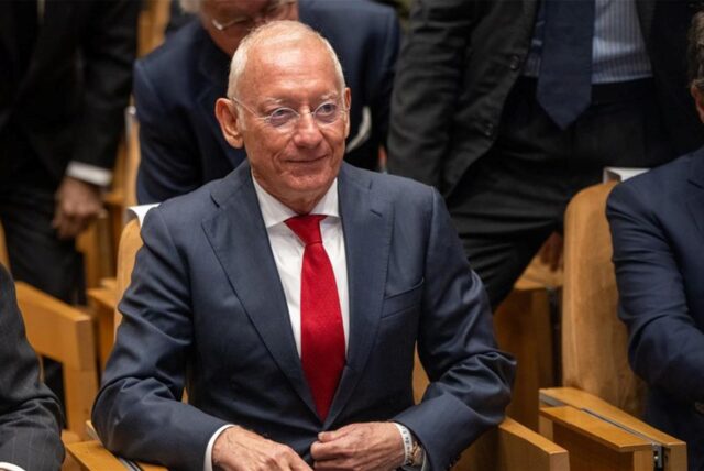 Isak Andic sitzt in einem Anzug auf einem Stuhl, um ihn herum weiter Männer in Anzügen.