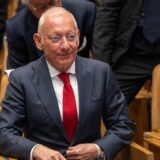 Isak Andic sitzt in einem Anzug auf einem Stuhl, um ihn herum weiter Männer in Anzügen.