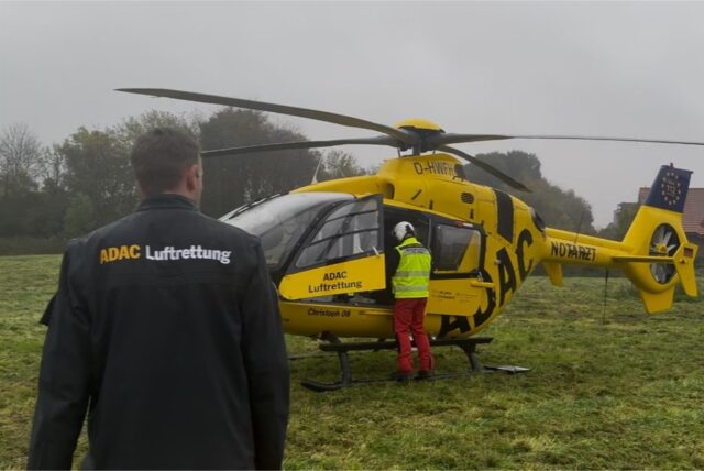 Ein Mann mit Jacke geht auf gelben Hubschrauber zu. Ein Mann mit Helm und gelber Weste steht am Hubschrauber.