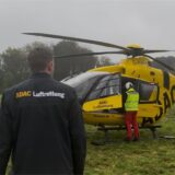 Ein Mann mit Jacke geht auf gelben Hubschrauber zu. Ein Mann mit Helm und gelber Weste steht am Hubschrauber.